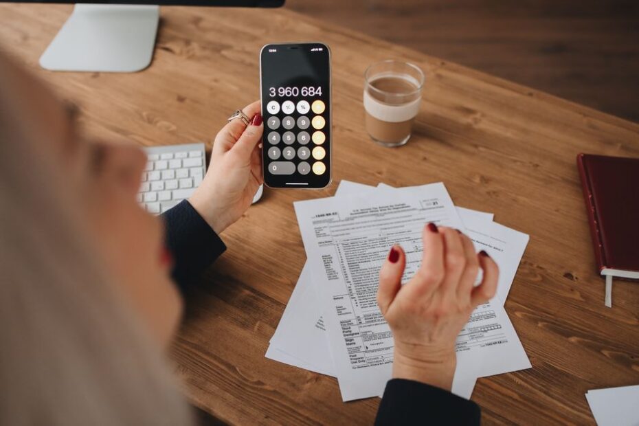 A woman calculates on her smartphone while working with documents at a desk.