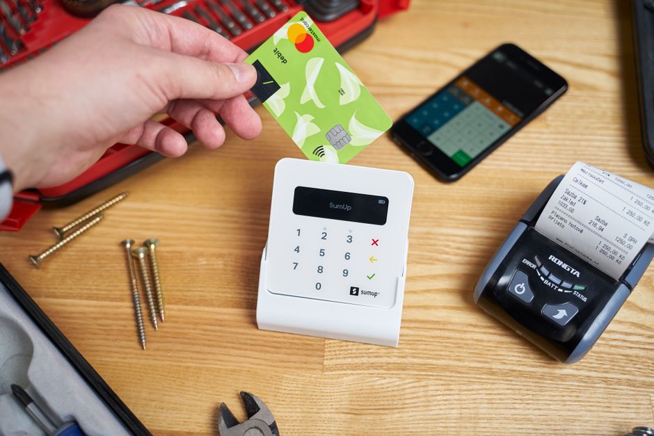 Faceless customer paying by small bright plastic card while using cash register among smartphone and tools on wooden table in bright studio